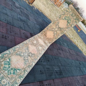 Peut inclure: Un tapis long et rectangulaire à l'aspect délavé et vintage. Le tapis a un fond rose clair avec un motif floral vert. Le tapis est posé sur une surface carrelée noire et brune.