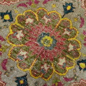 May include: A woven rug featuring a large central floral medallion in shades of red, olive green, and yellow, set against a light brown background. The design includes intricate geometric patterns, small teal accents, and pink floral motifs.