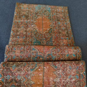 May include: A vintage Turkish runner rug with a brown, red, and teal geometric pattern. The rug is folded in thirds, showing the intricate details of the design.