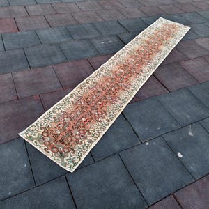 Pode incluir: Um tapete longo e retangular com um intrincado padrão floral em tons de vermelho, laranja e creme. O tapete tem um aspeto desgastado e vintage, com uma borda creme e um design central com motivos semelhantes a paisley. O tapete está colocado numa superfície cinza escuro e castanho avermelhado.