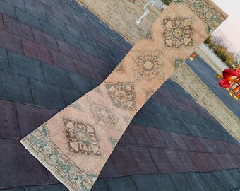 May include: A long, rectangular rug with a light pink background and a repeating pattern of green, brown, and white floral designs. The rug is folded in half and lying on a black and gray tiled surface.