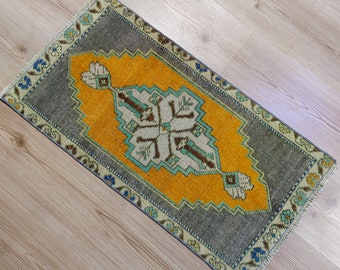 Puede incluir: Una pequeña alfombra rectangular con un diseño central amarillo enmarcado por un borde gris. La alfombra presenta un patrón simétrico detallado en tonos de blanco, turquesa y marrón. Los bordes están acabados con un borde decorativo azul y beige.