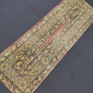Puede incluir: Una alfombra rectangular y desgastada con un aspecto vintage descolorido. La alfombra presenta un diseño central con patrones florales y geométricos en beige, rojo y negro. Dimensiones: aproximadamente 60 x 180 cm.