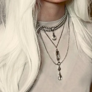 May include: A layered silver necklace set. The necklaces feature various chain styles, including beaded and delicate link chains. Each necklace has a different pendant, including teardrop and rectangular shapes. The jewelry is worn against a white background.