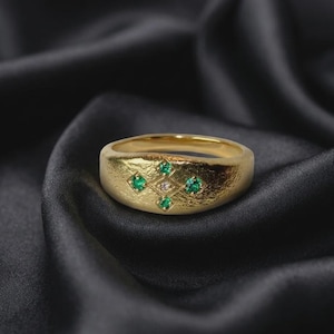 May include: A gold ring with a textured, hammered surface featuring a central diamond-shaped engraving. Four small green gemstones are set at the points of the diamond, with a single clear, colourless gemstone positioned in the very centre.