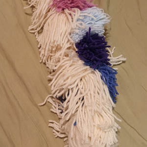 Könnte beinhalten: Ein langer, weißer Garnpompon mit rosa, hellblauen, dunkelblauen und violetten Garnpompons am Ende.