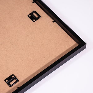 K&ouml;nnte beinhalten: Nahaufnahme der R&uuml;ckseite eines schwarzen Bilderrahmens mit braunem Kartonr&uuml;cken. Zwei Metall-Aufh&auml;nger sind auf der R&uuml;ckseite des Rahmens sichtbar.