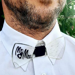 Peut inclure: Un nœud papillon blanc avec les mots "Mr & Mrs" imprimés en noir. Le nœud papillon est fait d'un matériau transparent et a un ruban noir.