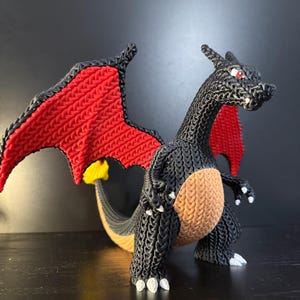 May include: A dragon-like figure constructed from small black, red, tan, and yellow rubber bands. The creature features large red wings, a tan belly, white claws, and a yellow flame at the tip of its tail. The body has a textured, woven appearance.