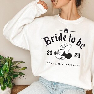 Puede incluir: Sudadera blanca con un gráfico negro de Mickey Mouse con el texto "Bride to be 20 24 Anaheim, California" impreso en ella.