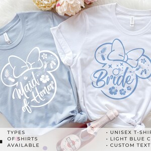 Può includere: Due magliette unisex, una azzurra e una bianca, con un design ispirato a Minnie Mouse. La maglietta azzurra dice "Maid of Honor", e la maglietta bianca dice "Bride". Entrambe le magliette hanno un fiocco ed elementi tropicali. L'immagine include anche testo sui tipi di magliette e il colore.