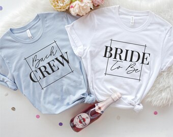 May include: Two short-sleeved t-shirts, one light blue and one white, with black text. The light blue shirt says "Bach Crew" and the white shirt says "Bride To Be". A bottle of sparkling wine is in the foreground.