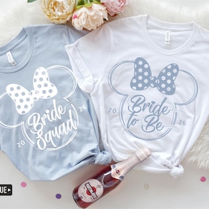 Puede incluir: Dos camisetas, una azul bebé y otra blanca, con un diseño inspirado en Minnie Mouse. La camiseta azul bebé dice "Bride Squad 26" y la blanca "Bride to Be 26". Una botella de vino espumoso Martini Rossi en primer plano.