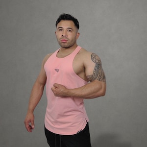 May include: A light pink tank top with a small logo on the chest, paired with black athletic shorts. The tank top has a racerback design. The shorts have a drawstring and a small logo near the hem. The background is a solid gray.