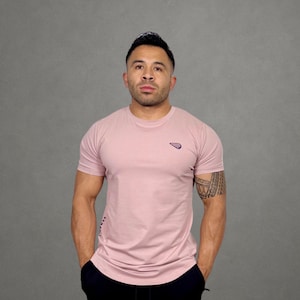 May include: A light pink crew neck t-shirt with a small logo on the chest. The shirt is paired with black shorts. The model is standing in front of a gray background.