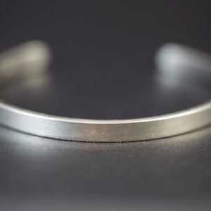 Puede incluir: Primer plano de una pulsera de plata. La pulsera tiene un dise&ntilde;o minimalista con una banda plana y rectangular. El metal tiene un acabado cepillado, lo que le da un aspecto mate. Los extremos est&aacute;n abiertos.