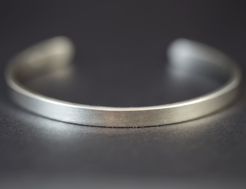 Puede incluir: Primer plano de una pulsera de plata. La pulsera tiene un dise&ntilde;o minimalista con una banda plana y rectangular. El metal tiene un acabado cepillado, lo que le da un aspecto mate. Los extremos est&aacute;n abiertos.