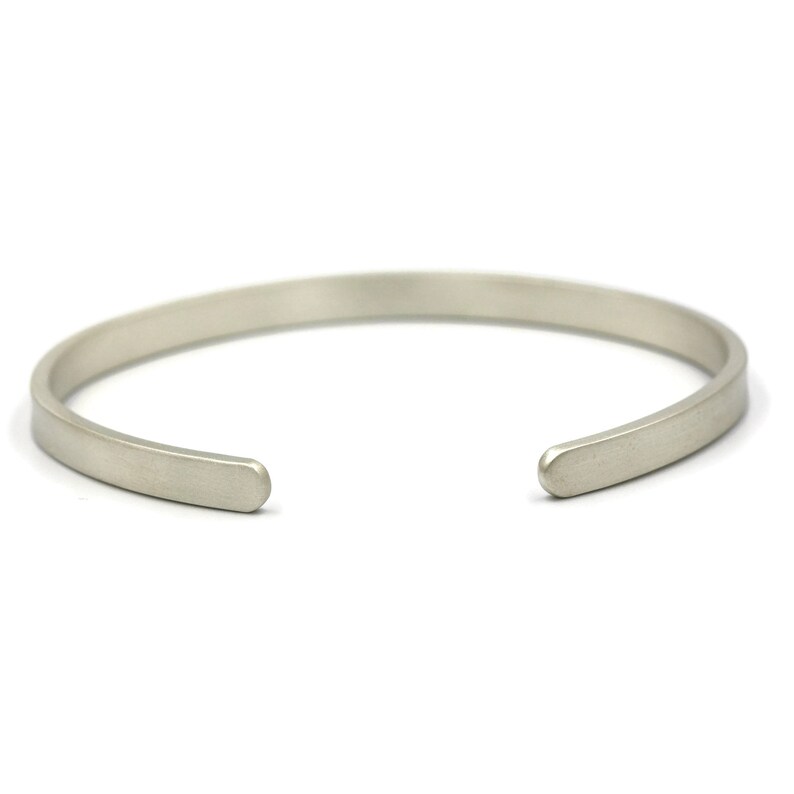 Puede incluir: Una pulsera de pu&ntilde;o plateada con un dise&ntilde;o minimalista. La pulsera abierta tiene un acabado liso y mate y una forma rectangular redondeada. La pulsera se muestra sobre un fondo blanco.