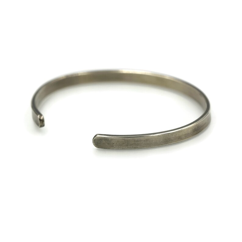 Puede incluir: Una pulsera de pu&ntilde;o de color plateado con un dise&ntilde;o abierto y sencillo. La pulsera tiene un acabado liso y mate y una forma ligeramente redondeada. Los extremos abiertos permiten un f&aacute;cil ajuste. La pulsera se muestra sobre un fondo blanco.