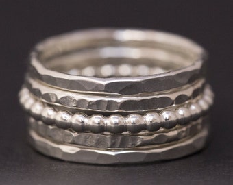 May include: Five silver rings stacked together. The top ring has a beaded design, while the other rings have a hammered texture.