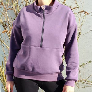 Puede incluir: Una sudadera morada con cremallera y bolsillo canguro. La sudadera tiene mangas largas y cuello alto.
