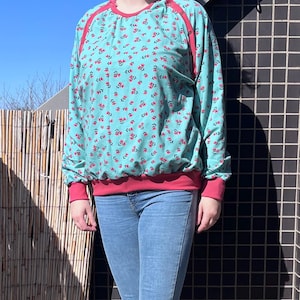 May include: A turquoise blue sweatshirt with a small pink floral print. The sweatshirt has a red ribbed neckline and cuffs. The model is wearing blue jeans.