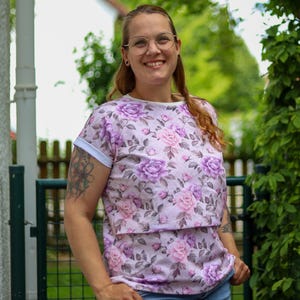 May include: A light pink floral print t-shirt with a round neckline and short sleeves. The shirt features a pattern of purple and pink roses. The sleeves have white cuffs. The person is wearing blue jeans.