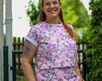 Könnte beinhalten: Hellrosa T-Shirt mit floralem Muster aus lila und rosa Rosen. Das Shirt hat einen Rundhalsausschnitt und kurze Ärmel mit weißen Manschetten. Die Person trägt eine blaue Jeans.