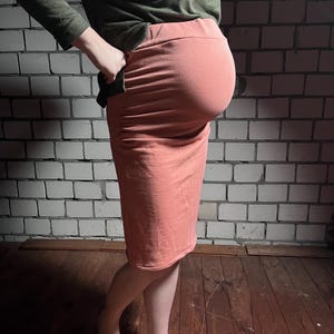 May include: A coral pencil skirt with a high waistband, worn with a long-sleeved olive green top. The skirt is knee-length and made of a soft, stretchy fabric. The outfit is simple and casual, suitable for everyday wear.