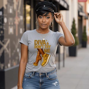 Könnte beinhalten: Graues T-Shirt mit dem Text "DIVA & DON'T TRY ME" in Leopardenmuster. Die Grafik zeigt Beine, die einen goldenen Plateau-Absatz und einen goldenen Sneaker tragen.