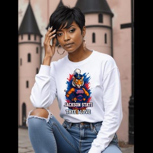 Puede incluir: Sudadera blanca de manga larga con un gráfico de tigre y el texto "JSU JACKSON STATE THEE I LOVE". El gráfico es azul, rojo y naranja. La sudadera se combina con unos vaqueros azules.