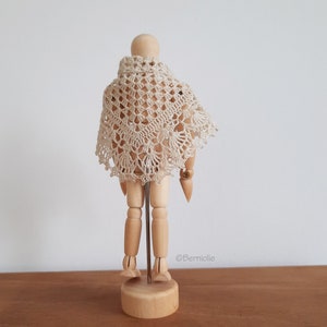 Puede incluir: Un chal de ganchillo color crema drapeado sobre un maniquí de madera. El chal presenta un diseño calado delicado con bordes festoneados. El maniquí está sobre una base de madera con un soporte de varilla metálica, sobre un fondo neutro.