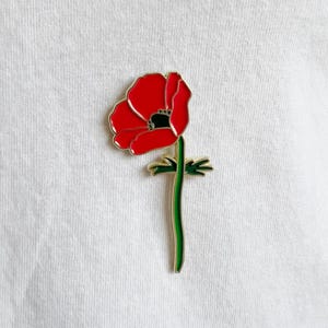 May include: A red poppy flower enamel pin with a gold outline and a green stem. The flower has a black center and is set against a white background. The pin is a decorative accessory.