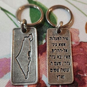 May include: Two rectangular silver-toned metal keychains. One keychain features an embossed map outline of Israel. The other keychain displays Hebrew text that reads: שיר למעלות אשא עיני אל-ההרים מאין יבא עזרי עזרי מעם ה' עושה שמים וארץ.