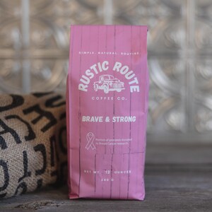May include: A pink bag of coffee from Rustic Route Coffee Co. with the words "Brave & Strong" and a pink ribbon symbol. The bag also states "Net wt. 12 ounces" and "340 g". A burlap sack with black text is to the left.