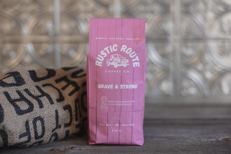 May include: A pink bag of coffee from Rustic Route Coffee Co. with the words "Brave & Strong" and a pink ribbon symbol. The bag also states "Net wt. 12 ounces" and "340 g". A burlap sack with black text is to the left.