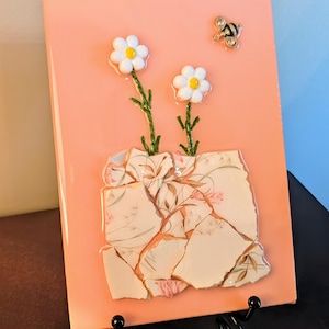 May include: A pink rectangular artwork features two white daisy flowers with yellow centers and a bee embellishment. The flowers have green stems and are above a mosaic of broken china with a floral pattern.