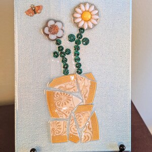 May include: A decorative canvas art piece featuring a light blue background with a mosaic design. The mosaic depicts two flowers with white petals and yellow centers, green stems, and a broken vase. An orange butterfly is also present.