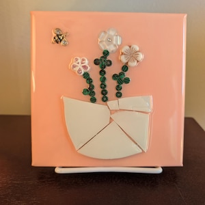 May include: A square, peach-colored artwork featuring a white vase with three flowers and a bee. The flowers have white and tan petals with green stems. The bee is gold and silver. The artwork is mounted on a white metal stand.