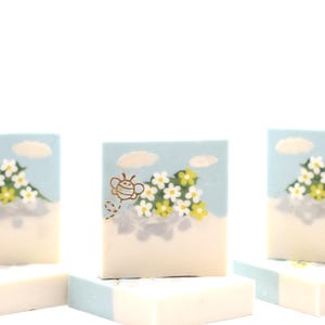 May include: Square soaps with a light blue top featuring a design of a bee, white and yellow flowers, and clouds. The soaps are stacked on top of each other, with a white and light blue color scheme.