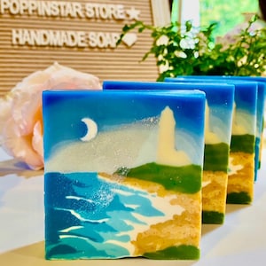 May include: Handmade soap bars with a coastal landscape design. Each bar features a blue sky, a white crescent moon, a lighthouse, and ocean waves in shades of blue and green. The soap has a sandy beach base.