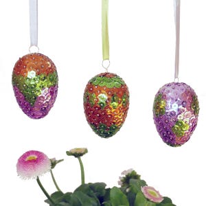 Könnte beinhalten: Drei dekorative Ostereier-Ornamente. Jedes Ei ist mit bunten Pailletten in Orange-, Grün- und Lilatönen bedeckt. Sie hängen an weißen und gelben Bändern. Eine kleine Topfpflanze mit rosa und weißen Blüten befindet sich im Vordergrund.