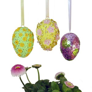 Könnte beinhalten: Drei dekorative Ostereier, die an Bändern hängen. Eines ist hellgrün mit gelben Blumen, ein anderes ist gelb mit rosa Blumen und Perlen, und das dritte ist lila mit Pailletten. Eine kleine Pflanze mit rosa und weißen Blüten befindet sich im Vordergrund.