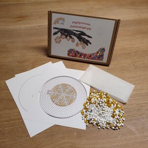 Könnte beinhalten: Ein Bastelset mit einem Karton mit Text und einem Weihnachtsbaum-Design. Das Set enthält weißes Papier, eine transparente Kunststoff-Steckplatte mit goldenem Design, einen Haufen weißer und goldener Perlen und ein weißes Rechteck.