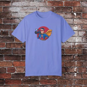 May include: A light blue t-shirt with a cartoon illustration of a superhero holding a camera. The superhero is wearing a blue and red costume with a yellow cape. The illustration is surrounded by a red circle.