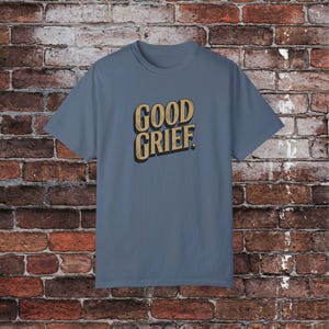 May include: A blue t-shirt with the text "GOOD GRIEF." printed in a vintage-style font in gold.