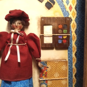 May include: A doll wearing a red dress and matching hat, positioned next to a wooden paint palette with six small colored squares and a white rectangular space. Below the palette is a patterned book with yellow, red, and blue floral designs.