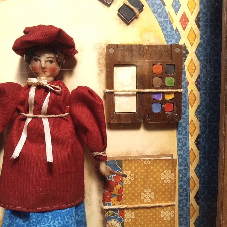 May include: A doll wearing a red dress and matching hat, positioned next to a wooden paint palette with six small colored squares and a white rectangular space. Below the palette is a patterned book with yellow, red, and blue floral designs.