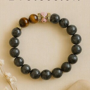 Può includere: Un braccialetto di perline con perline rotonde grigio scuro. Il braccialetto include una perla occhio di tigre e una perlina rosa e oro. Il testo spagnolo "Intención: Protección" è in alto.