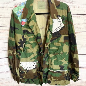 May include: A camouflage print jacket featuring a green, brown, and black pattern with added decorative elements. The jacket includes a light blue floral and green gingham patch on the shoulder, white lace trim on the pockets, and a white lace doily.